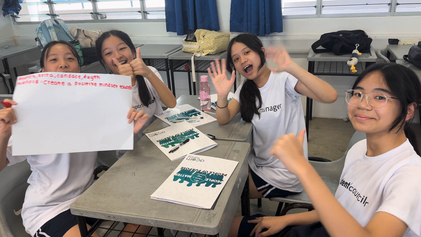 Four smiling teenage girls in white shirts sit at school desks, one holding a paper about creating a positive mindset.