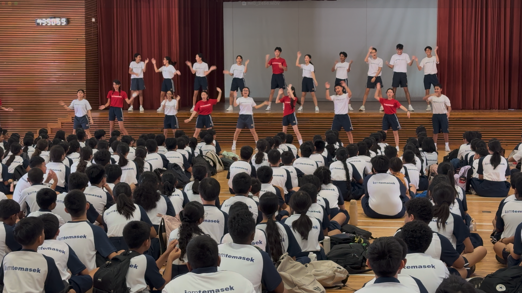 Students in "iamtemasek" shirts sit on a hall floor watching peers perform a dance on a tiered stage.