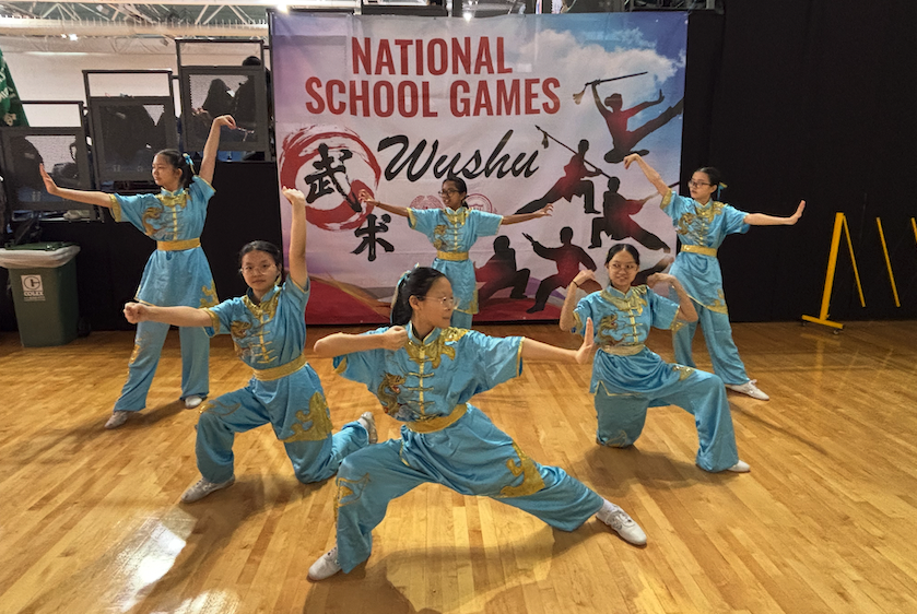 Six girls in blue and gold wushu uniforms perform synchronized martial arts poses at the National School Games Wushu event.