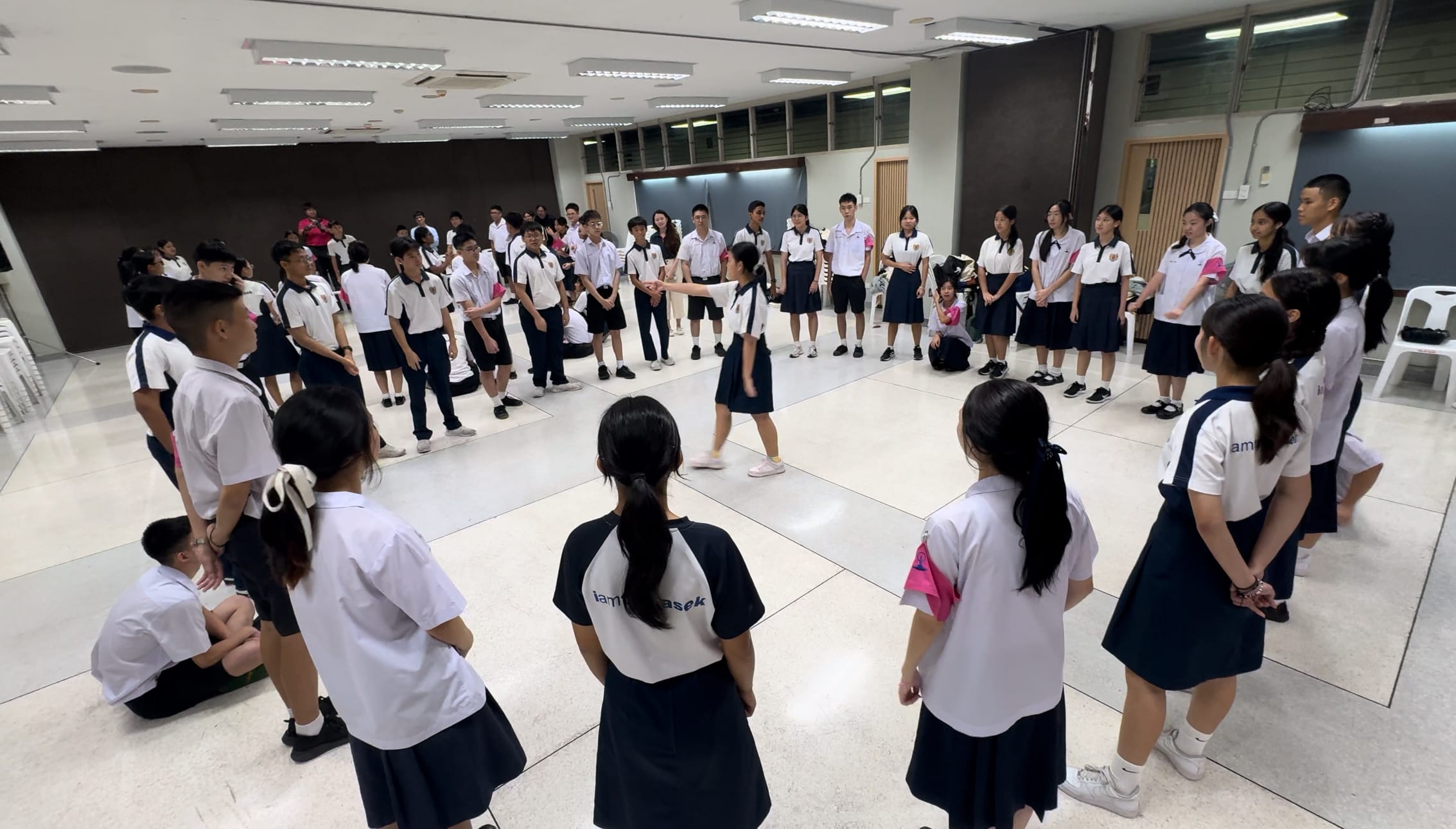 Students in white and navy uniforms stand in a large circle indoors while two students interact in the center.