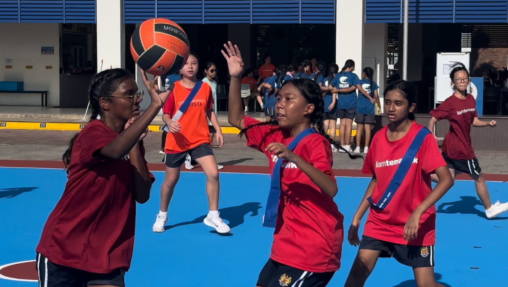 Girls in red jerseys play netball on a blue outdoor court, with one player holding a ball overhead.