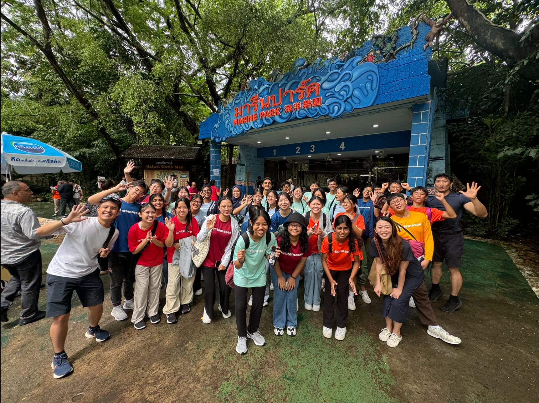 Large group of people posing and waving in front of a blue Marine Park entrance gate with Thai and Chinese text.
