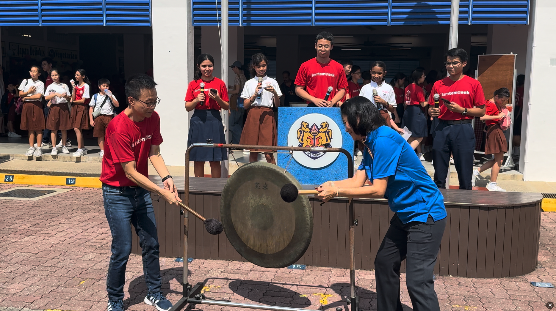 Two people strike a large gong outdoors at a school event while students in red shirts watch nearby.