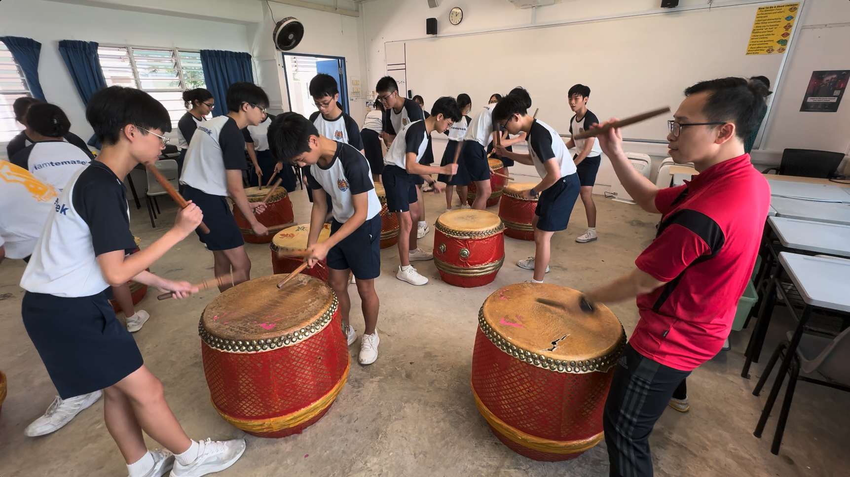Students in a classroom learning to play large red Chinese drums, guided by an instructor in a red shirt.