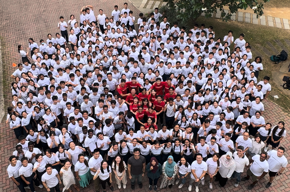 Large aerial group photo of hundreds of students mostly in white t-shirts, with a few in red at the center.