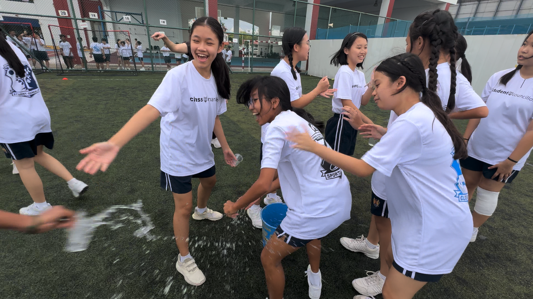 Girls in white school t-shirts laugh and play a water bucket game on a green sports field.