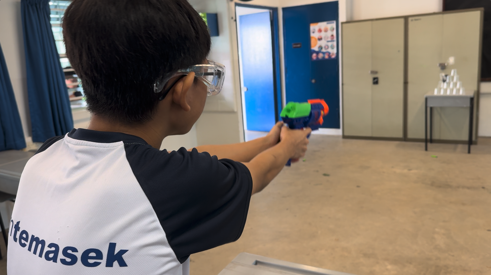 A boy wearing safety goggles and a Temasek shirt aims a colorful toy blaster toward a target indoors.