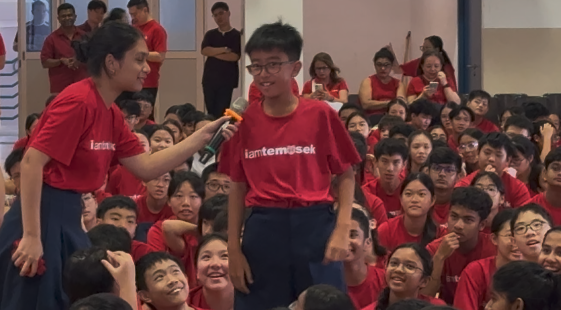 A student in a red "iamtemasek" shirt speaks into a microphone held by another student at a school assembly.