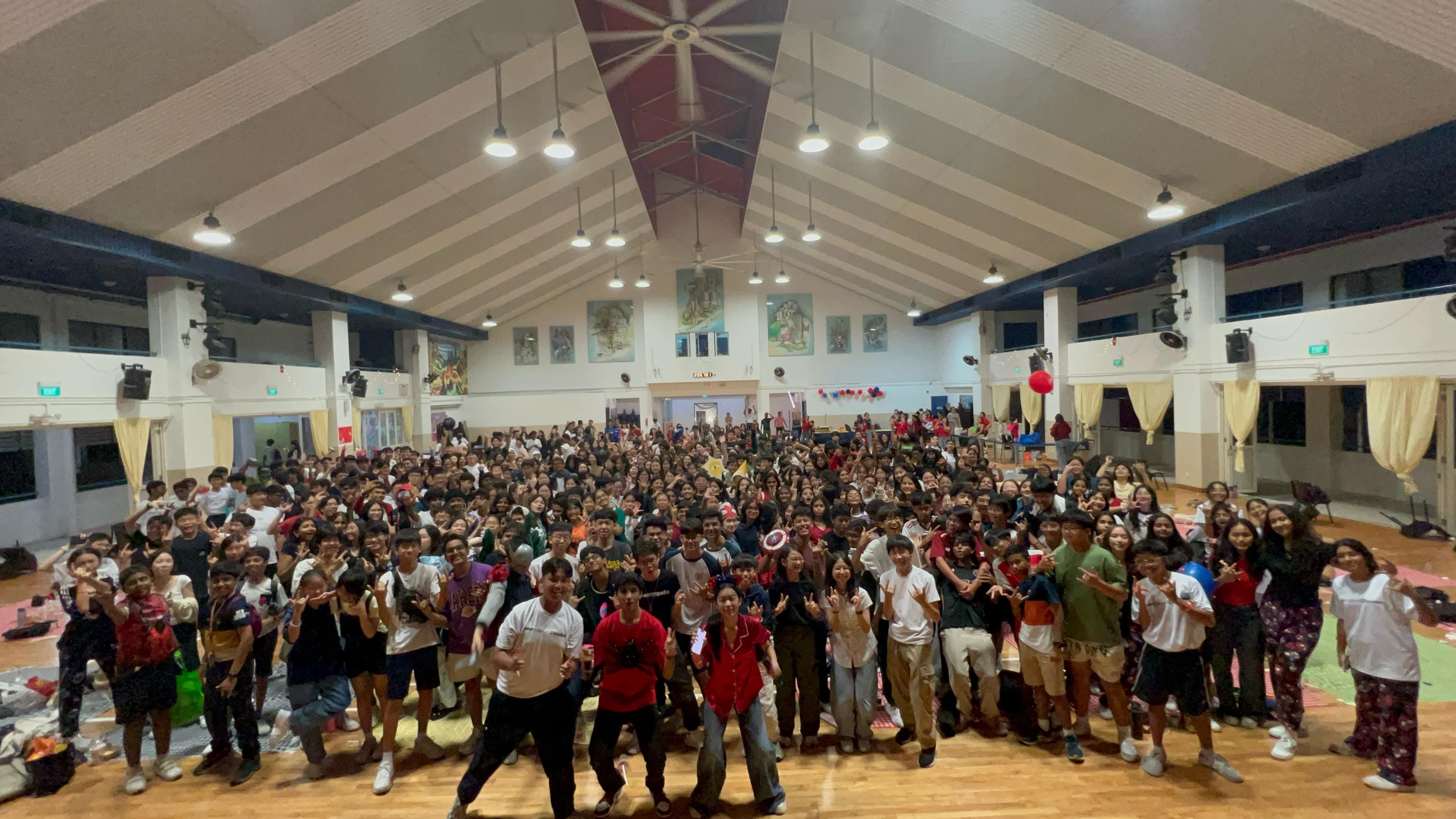 Large crowd of students posing and making hand signs inside a school hall with high vaulted ceiling.