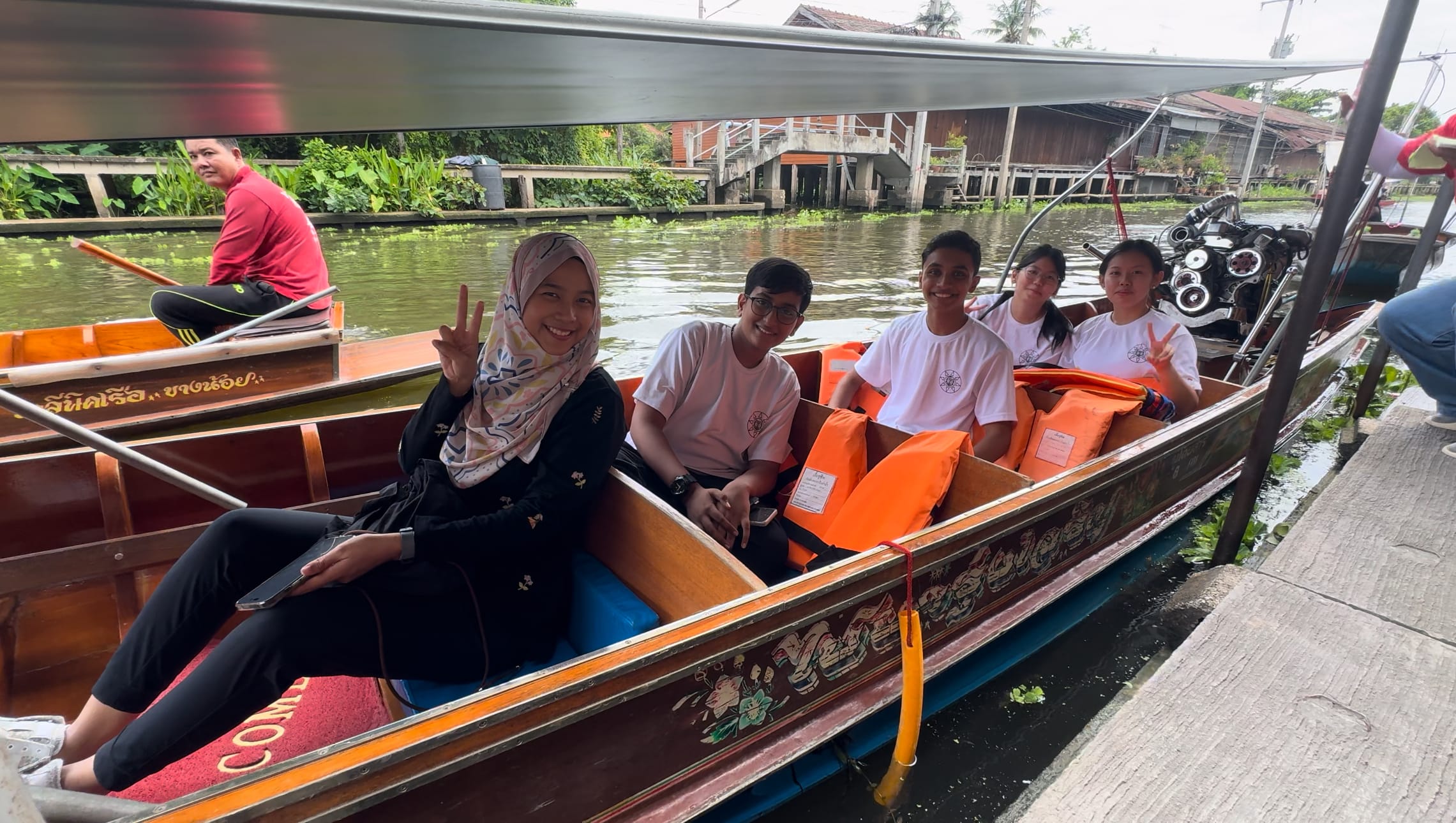 Five smiling people sit in a wooden canal boat with orange life jackets, posing with peace signs on a river.