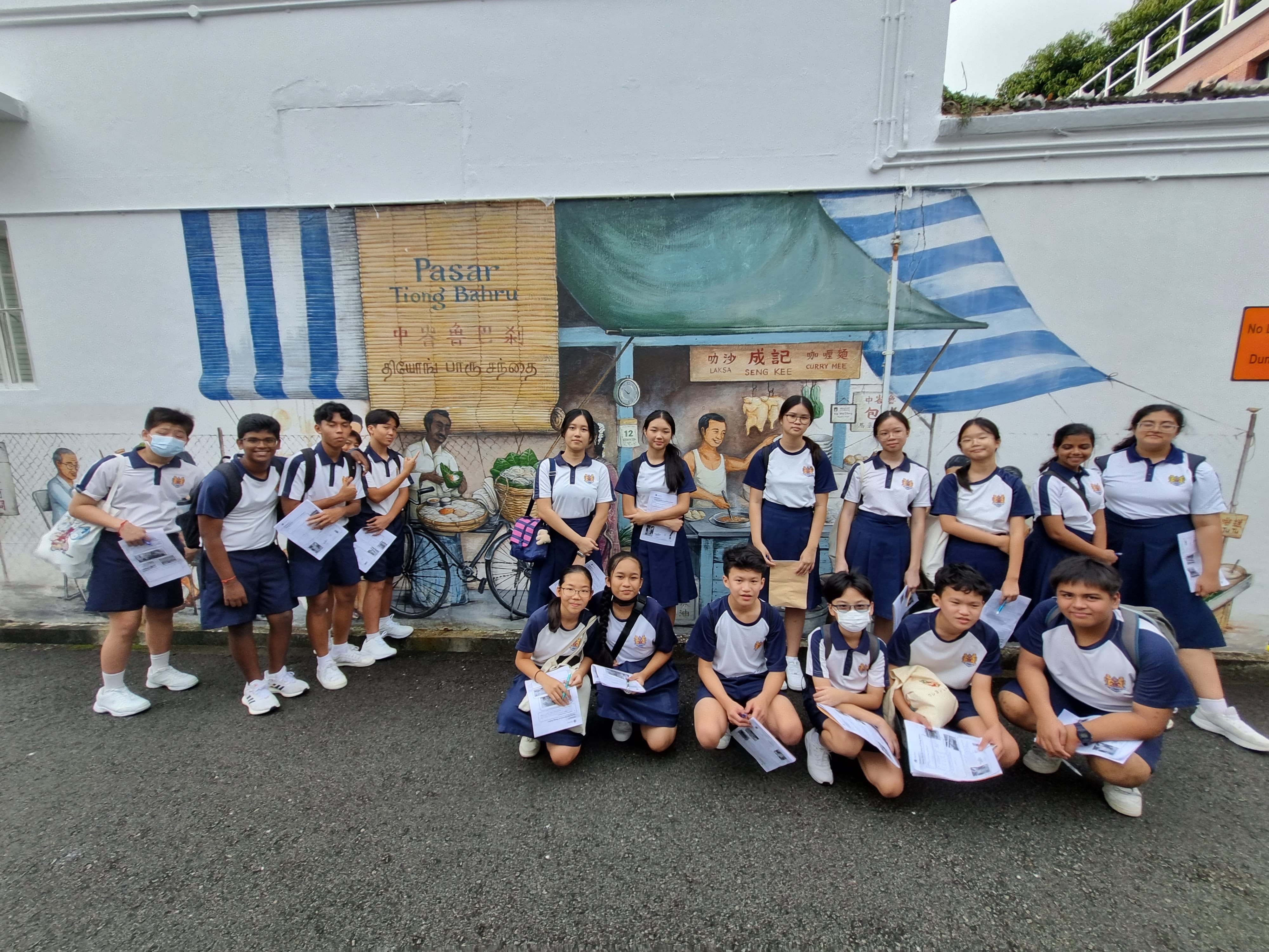 Students in navy and white uniforms pose in front of a Pasar Tiong Bahru street mural on a white wall.