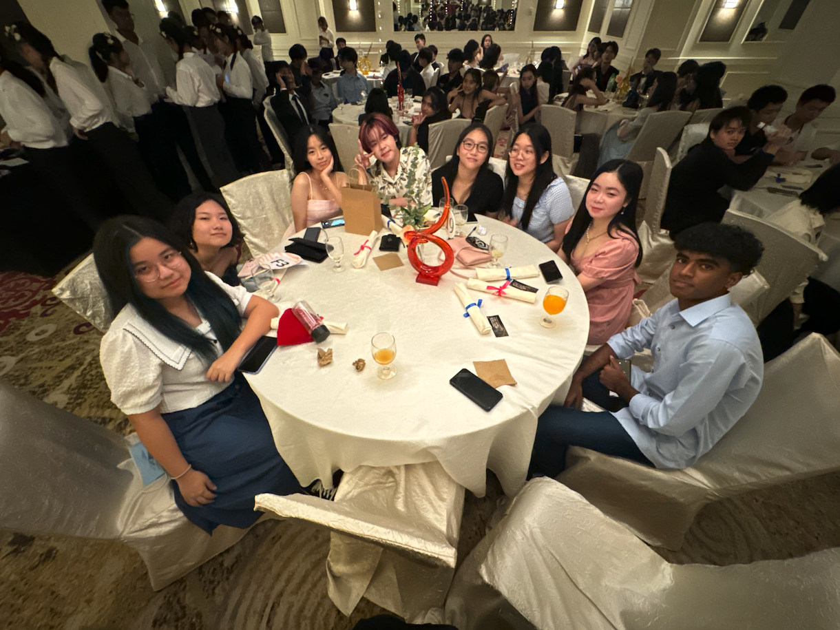A group of young people seated around a round white-clothed table in a large formal banquet hall.