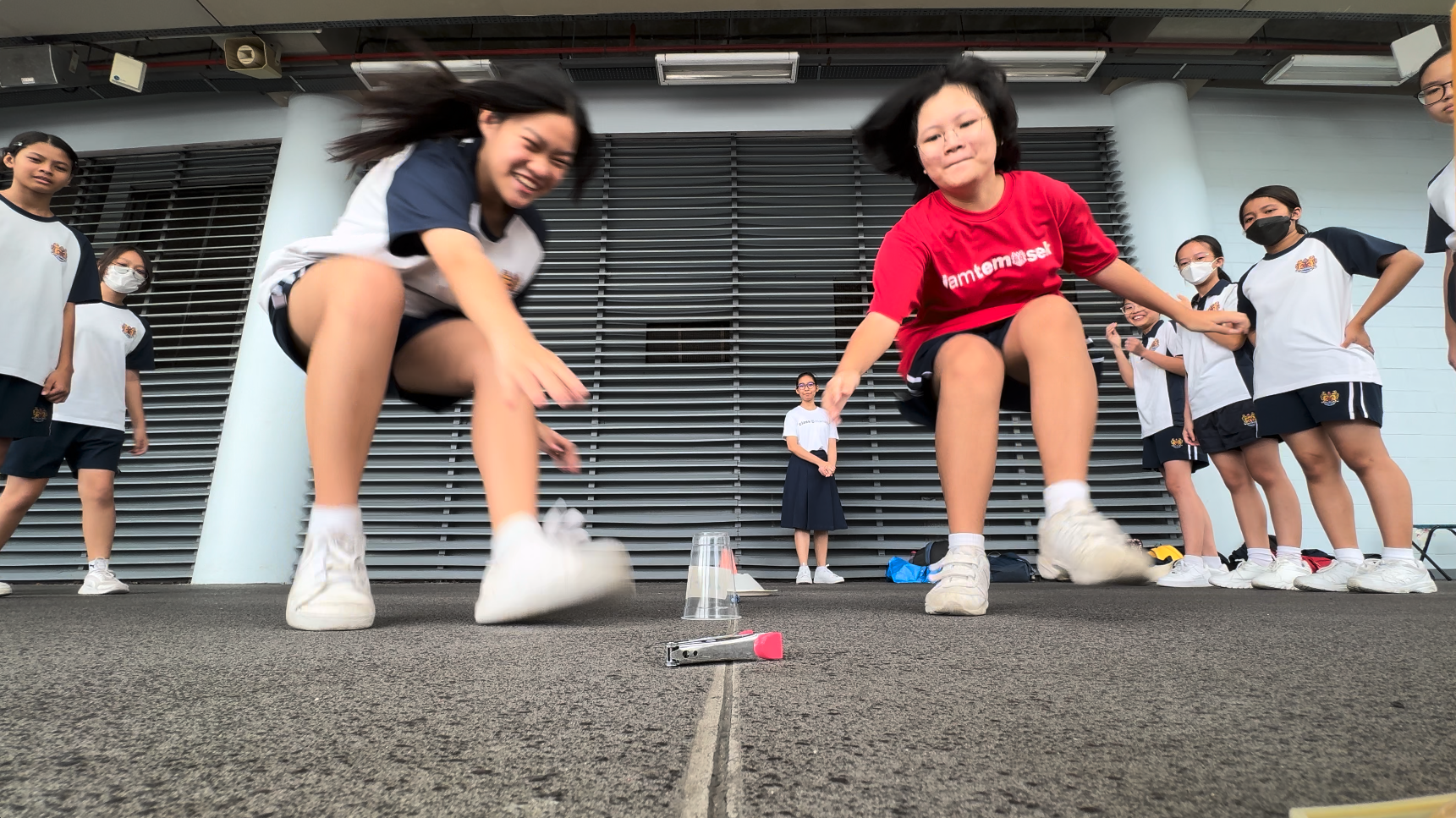 Two girls jump over a small cup on pavement while a group of students in school uniforms watches nearby.