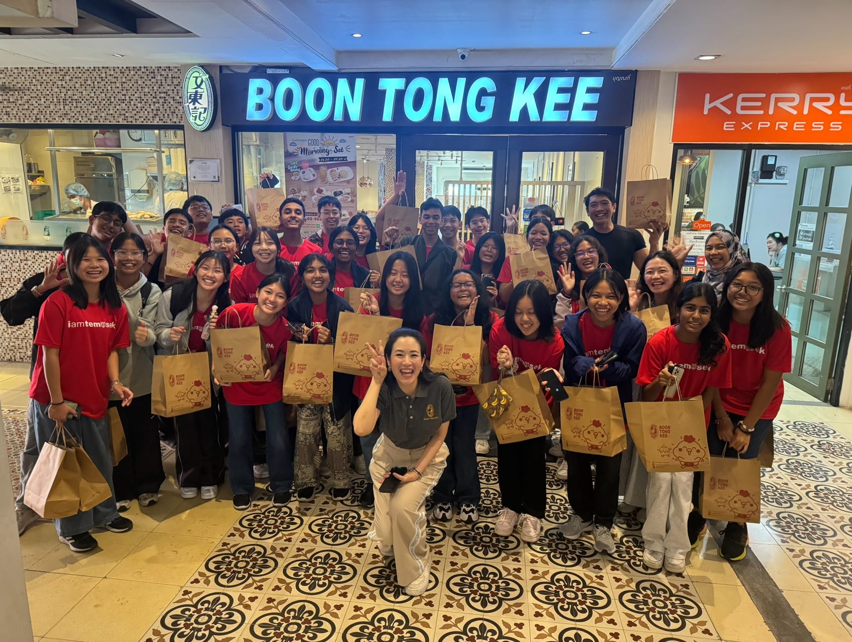 A large group in red shirts poses smiling outside Boon Tong Kee restaurant, holding branded paper bags.