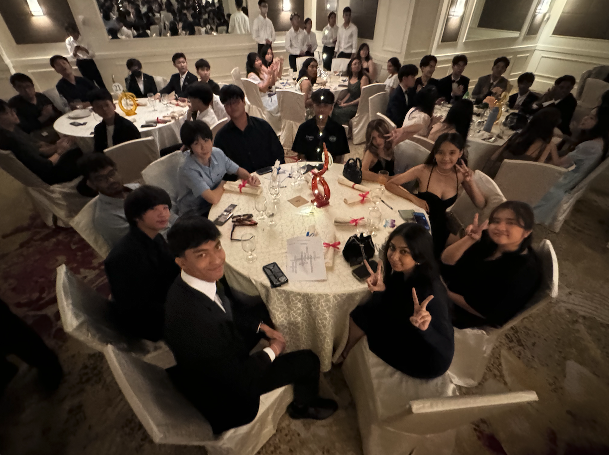 Young people in formal attire sit at round banquet tables in a large decorated hall, some posing with peace signs.