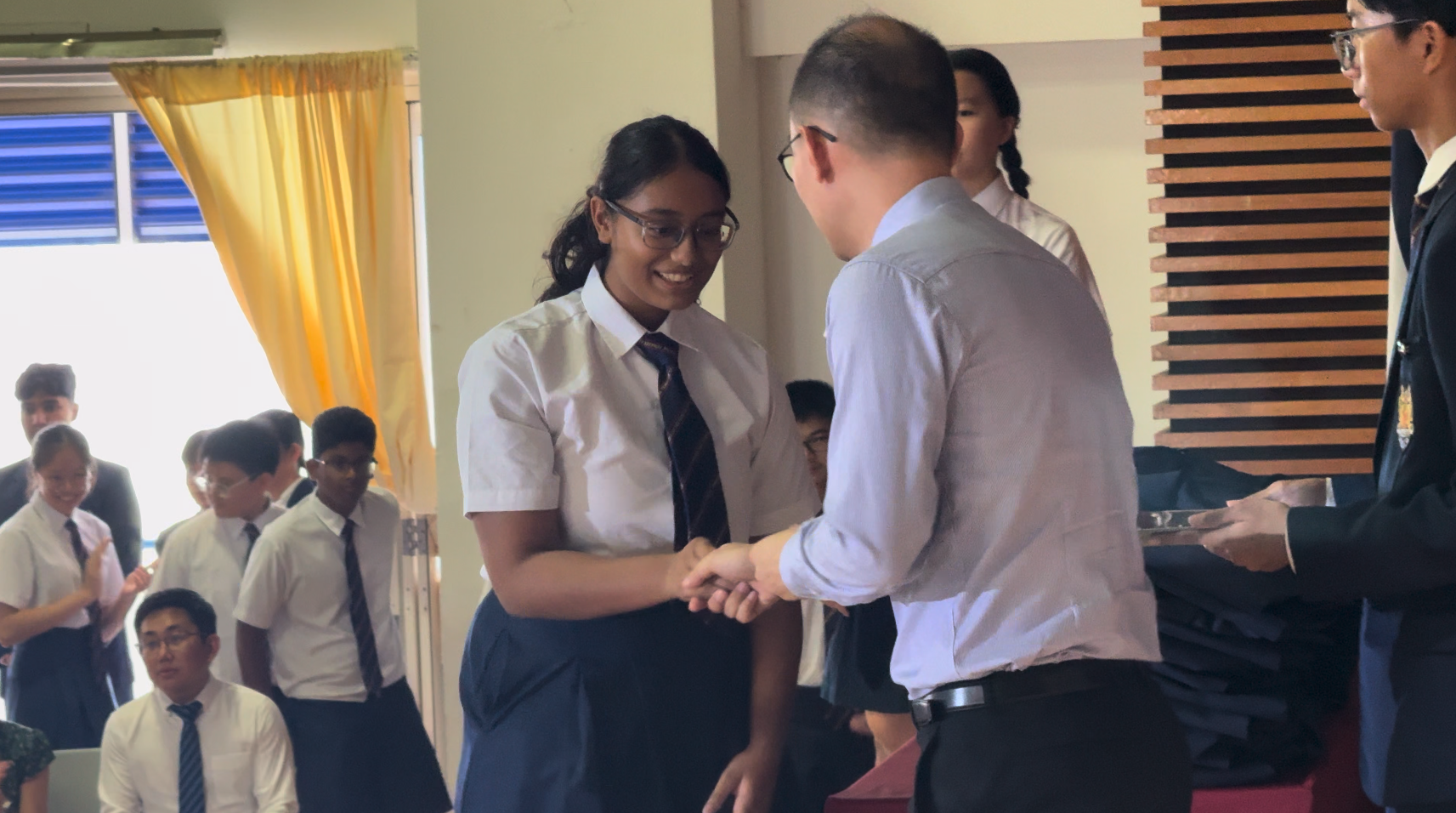 A smiling female student in uniform shakes hands with a man during a school award ceremony.
