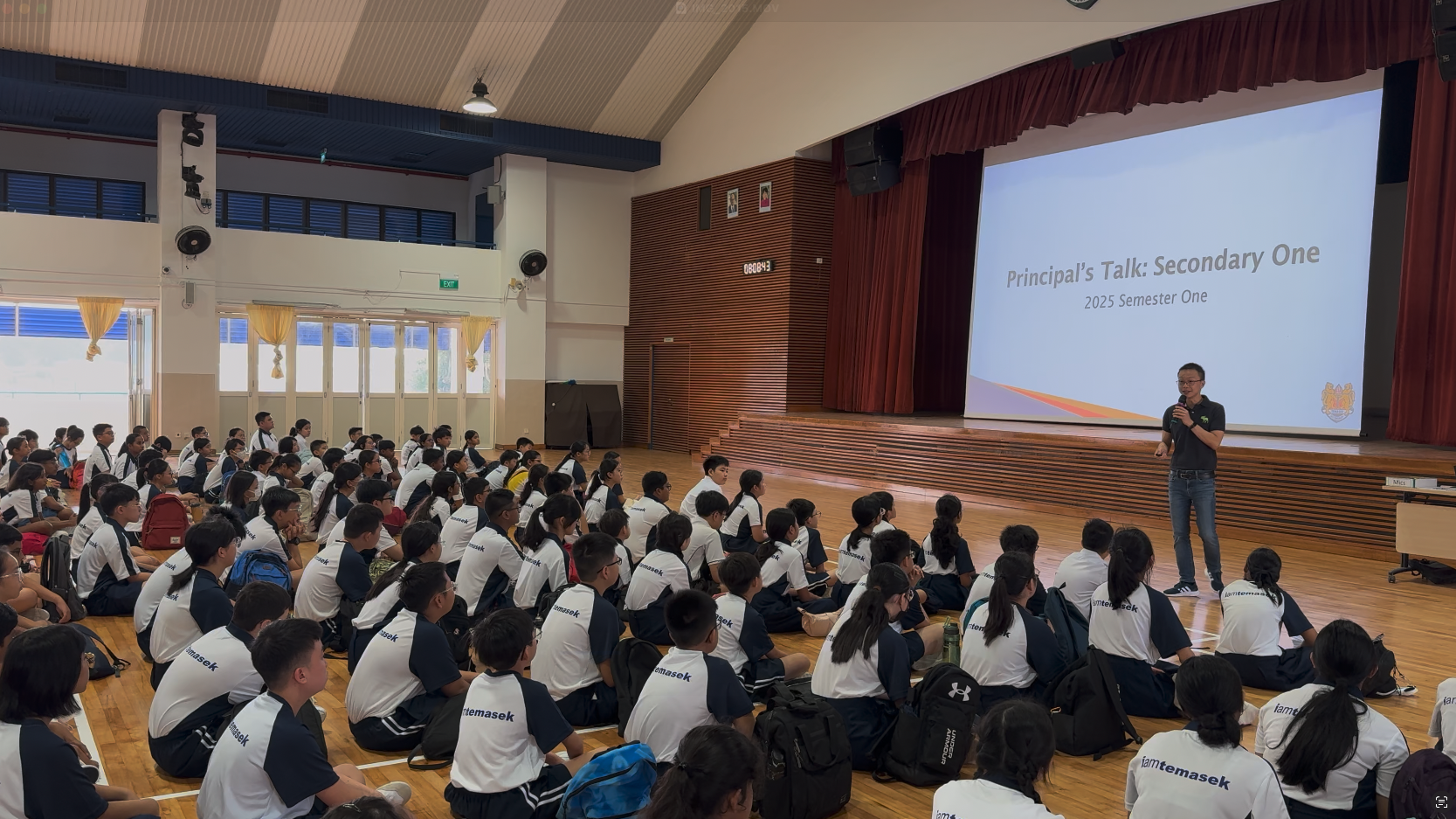 Students in school uniforms sit on a hall floor listening to a speaker presenting "Principal's Talk: Secondary One, 2025 Semester One."