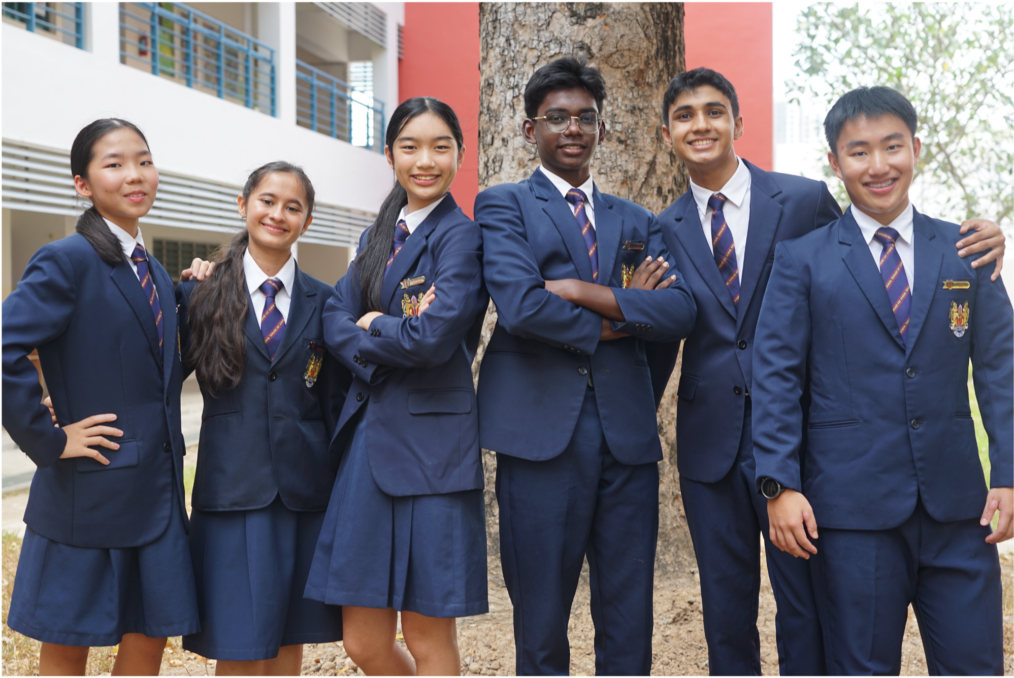 Six students in navy school uniforms pose smiling by a tree, arms crossed, with a campus building behind.