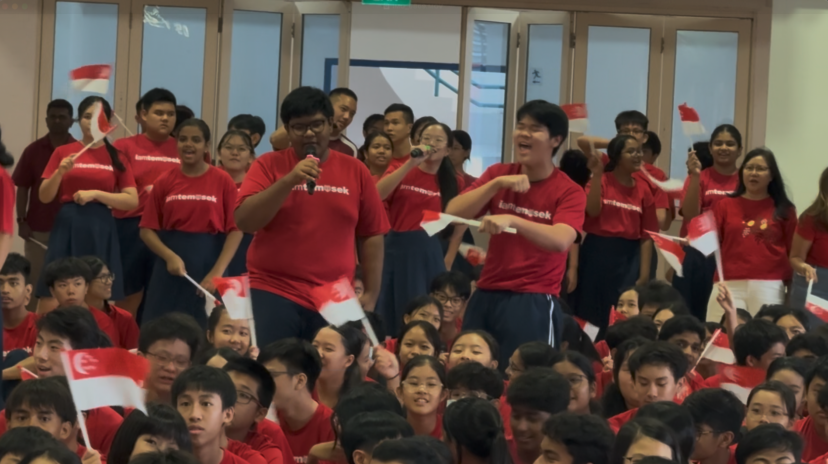 Students in red Temosek shirts wave Singapore flags and sing at a school celebration event.