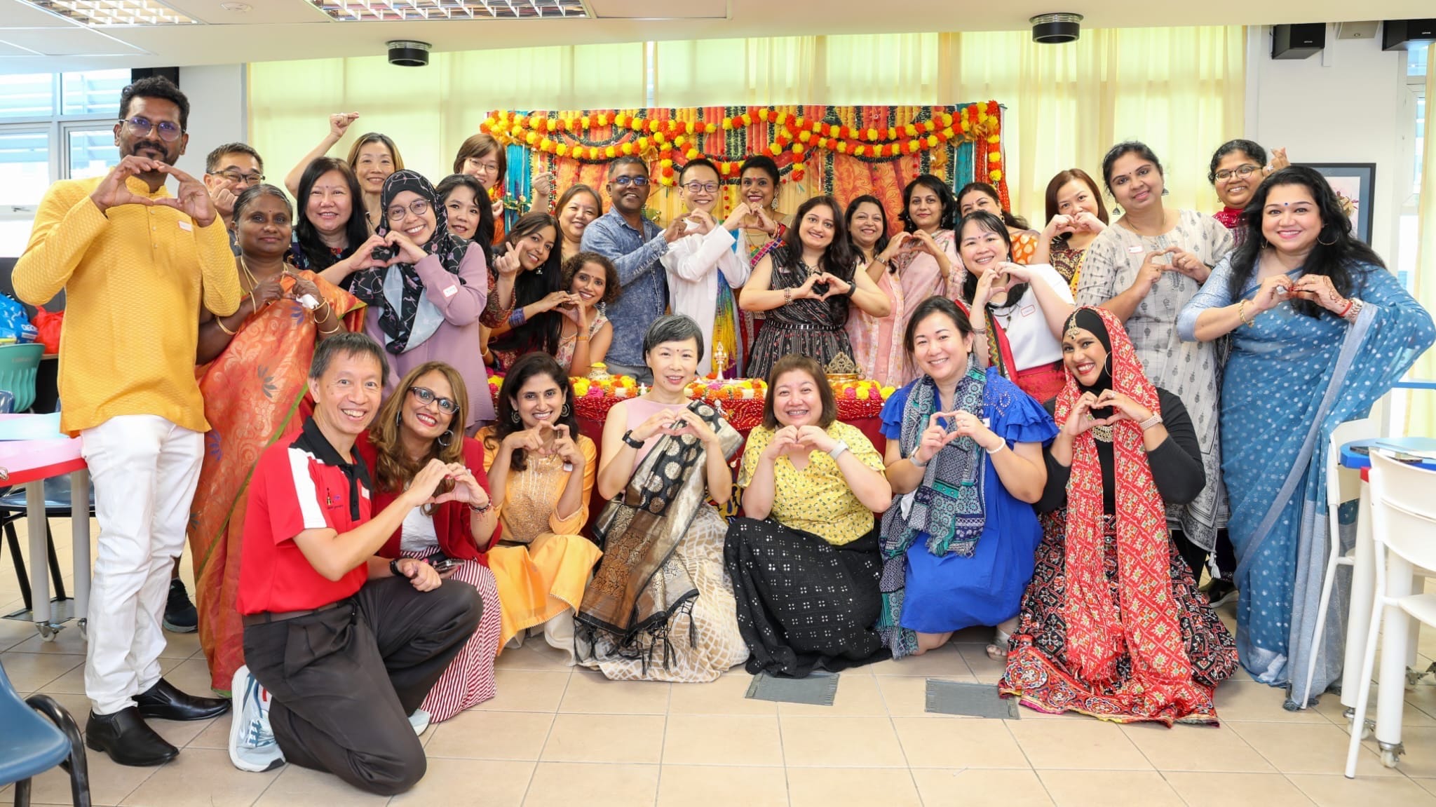 A large diverse group in colorful traditional attire poses making heart shapes with their hands before a festive floral backdrop.