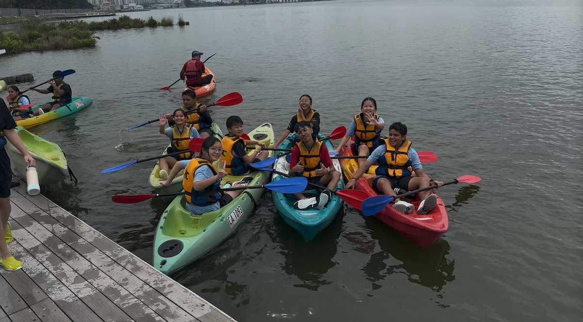 A group of young people in life jackets sit in colorful kayaks on calm water near a wooden dock.