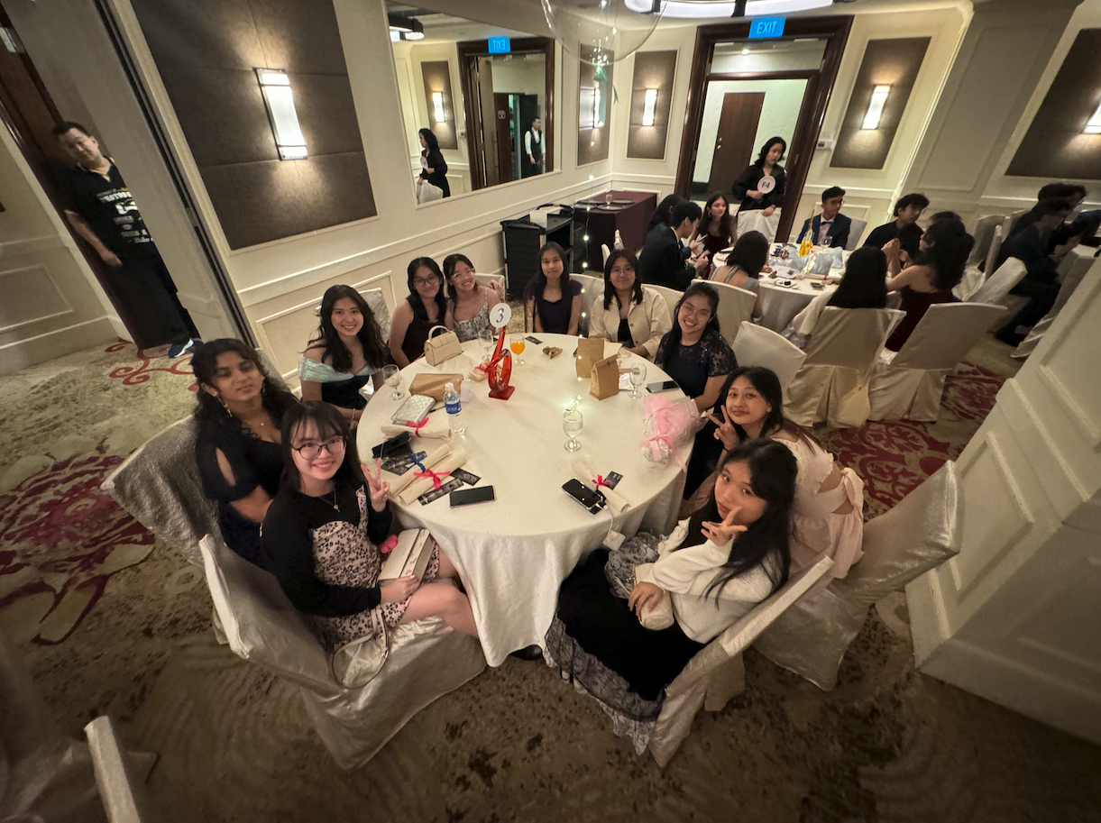 A group of young women smiling at a round banquet table in a formal dining hall during an event.