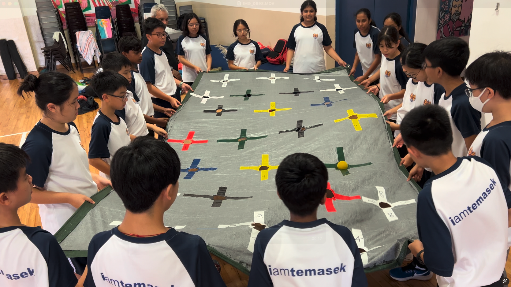 Students in Temasek uniforms stand around a large tarp covered with colorful paper airplanes during a group activity.