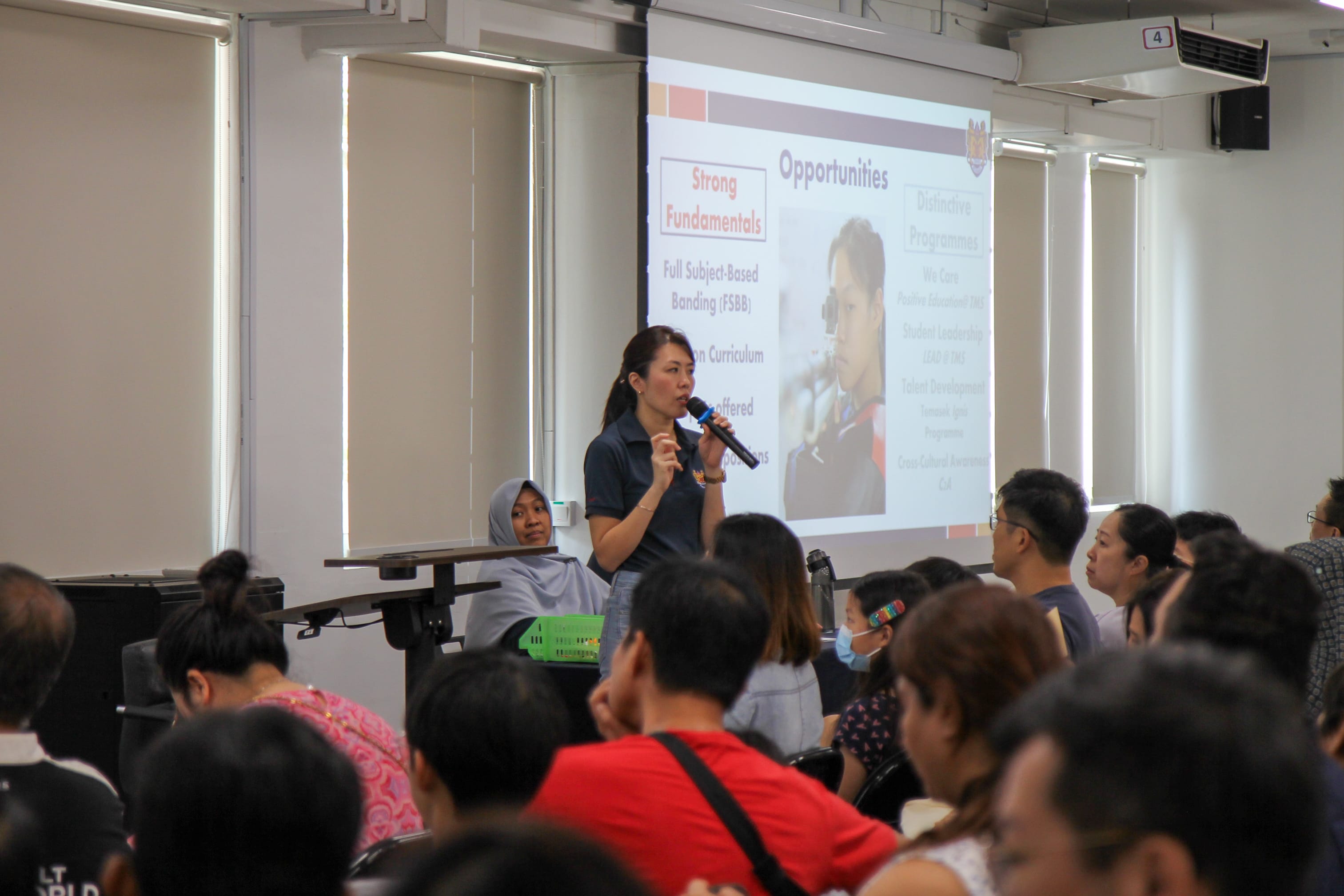 A woman speaks into a microphone before a seated audience with a projected slide showing "Strong Fundamentals" and "Opportunities."