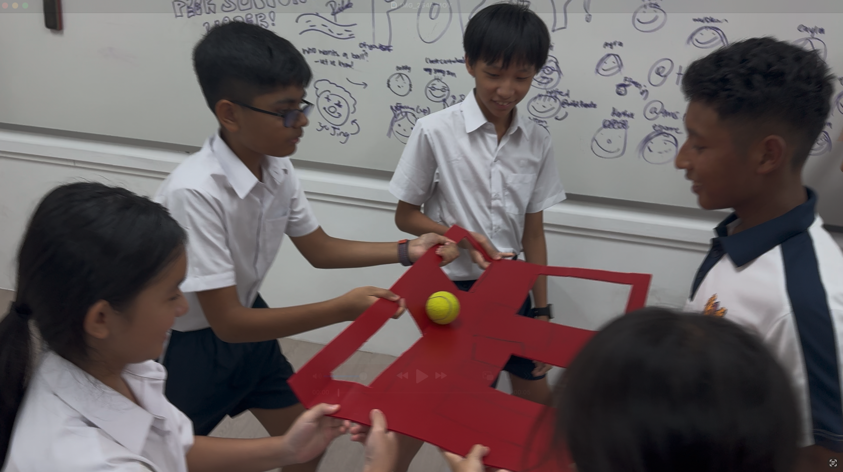 Students in school uniforms balance a yellow tennis ball on a red cardboard structure in a classroom.