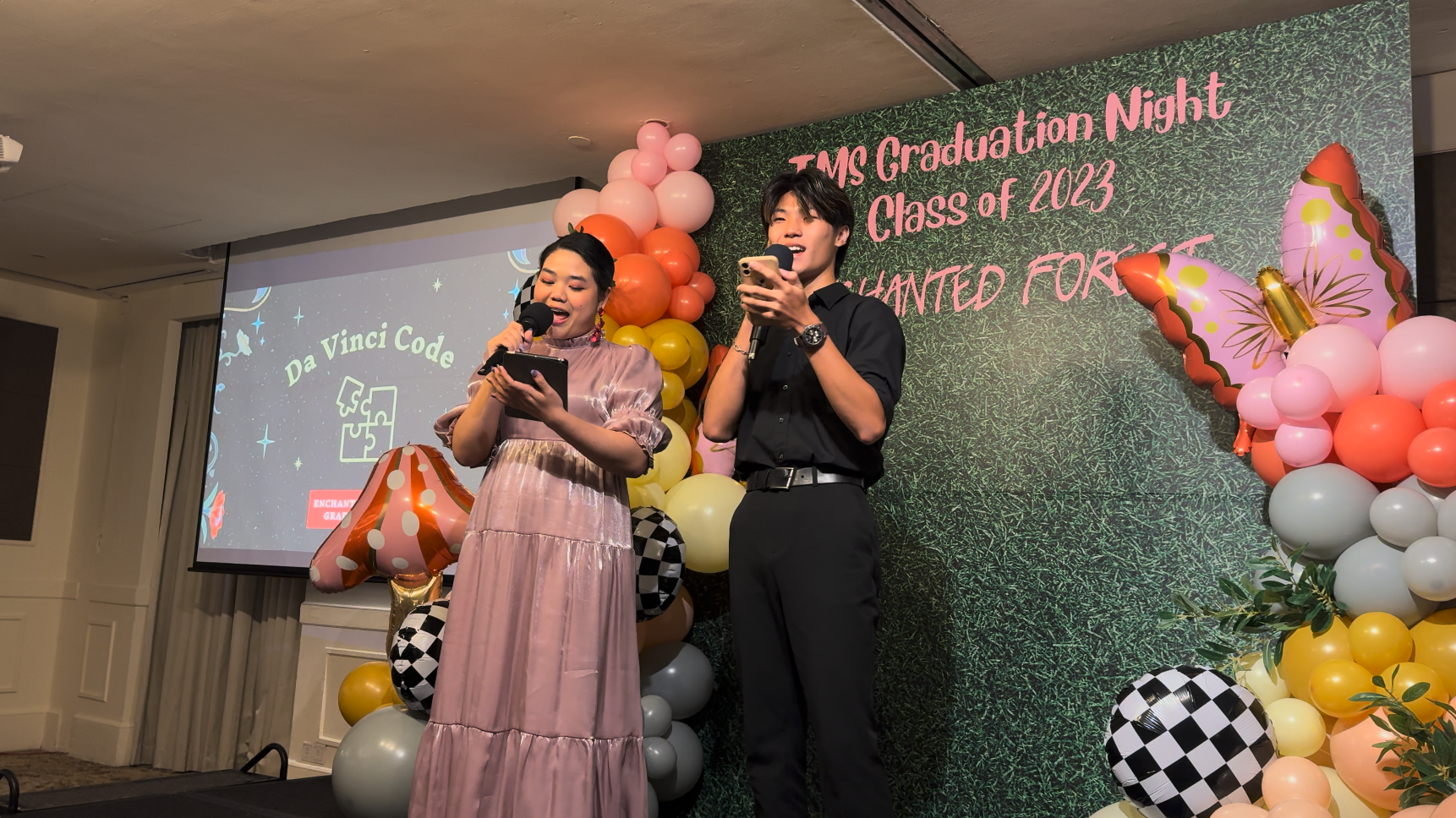 Two emcees hold microphones on stage at TMS Graduation Night Class of 2023, surrounded by colorful balloons.