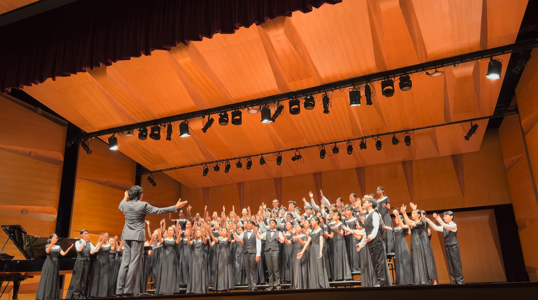 A conductor leads a large youth choir in gray formal attire performing on a lit concert hall stage.