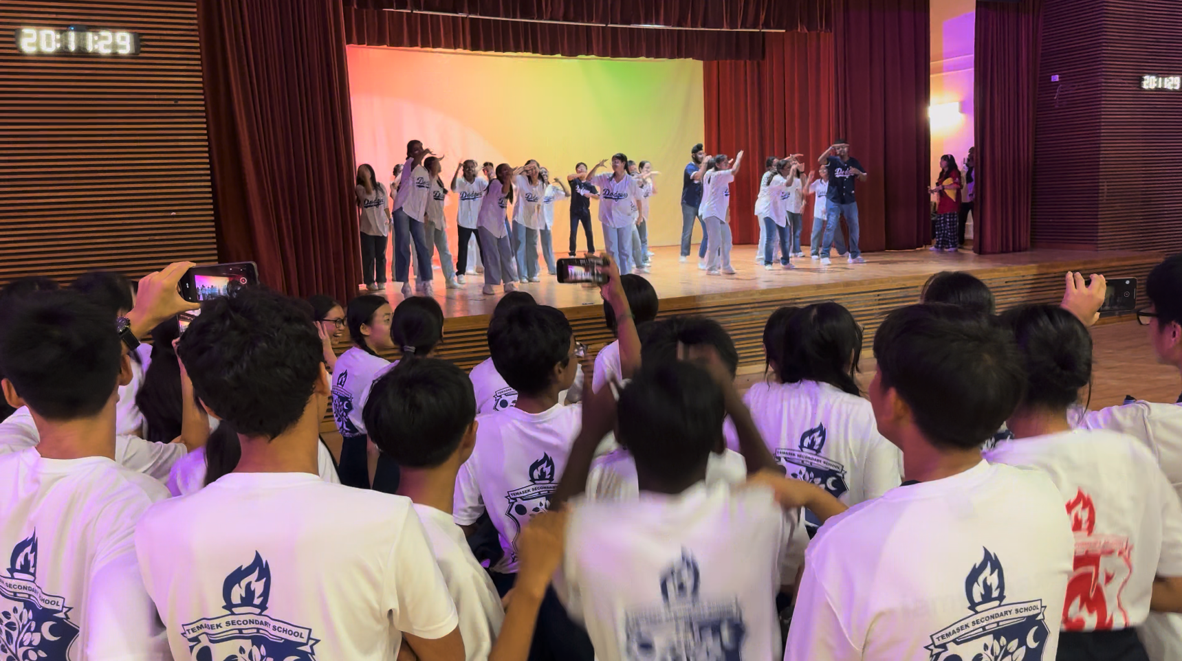 Students in Temasek Secondary School shirts watch and film a group of dancers performing on a lit stage.