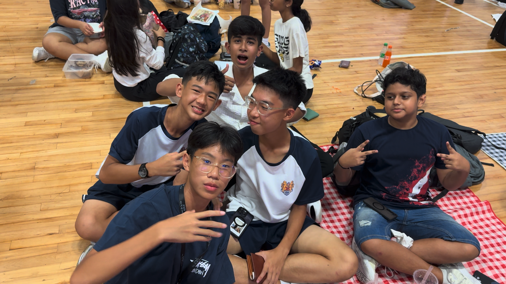 Five teenage boys sit on a gym floor taking a selfie, smiling and posing with hand gestures on a checkered blanket.