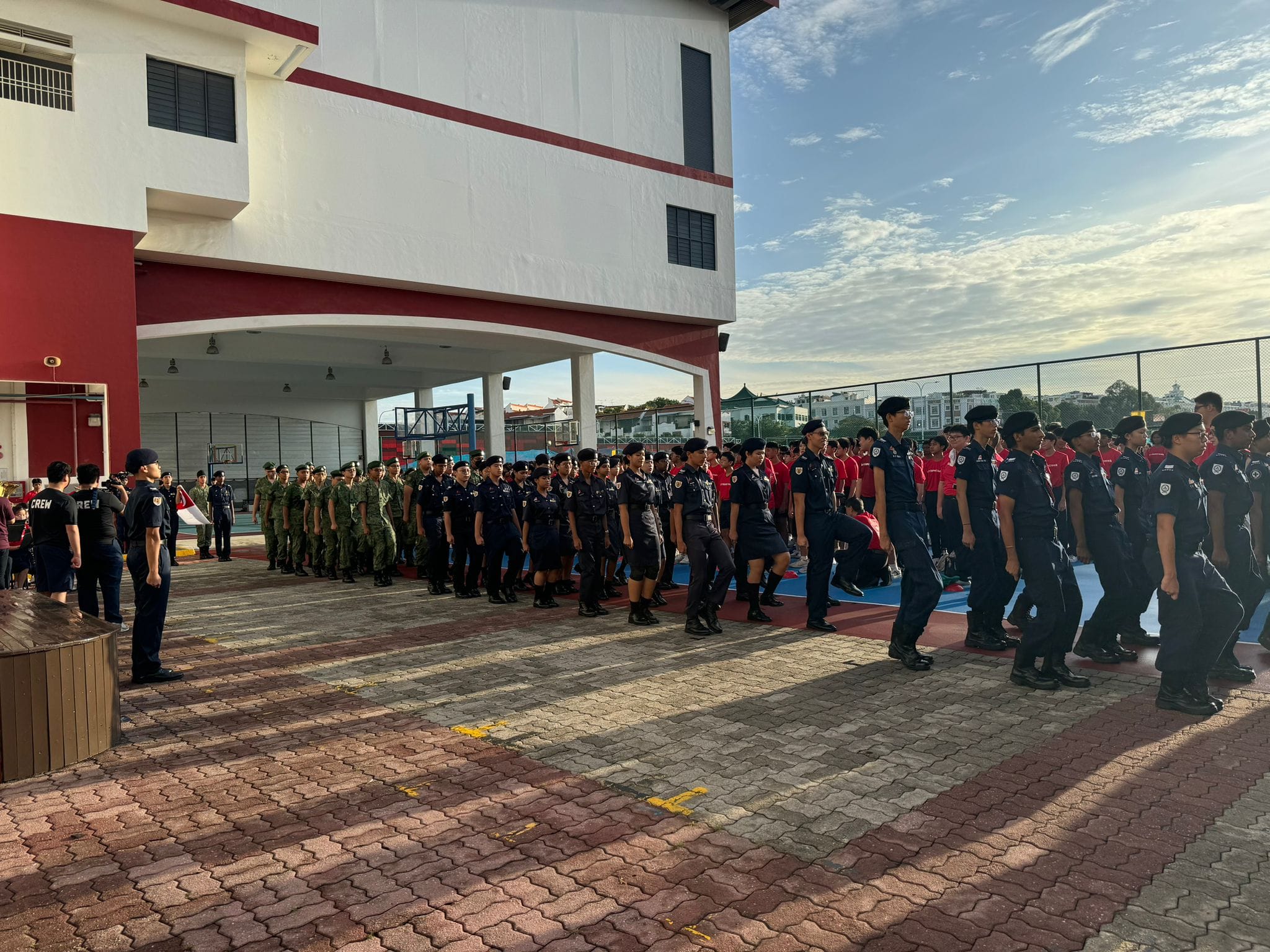 Students in military and police uniforms march in formation on a school courtyard beside a red and white building.