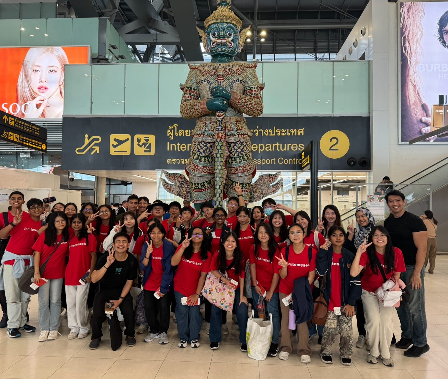 A large group in red Temasek shirts poses at a Thai airport's International Departures sign beneath a tall Yaksha guardian statue.