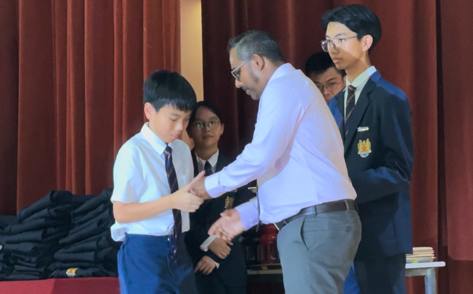 An adult man in a white shirt shakes hands with a young boy in school uniform on a stage with red curtains.