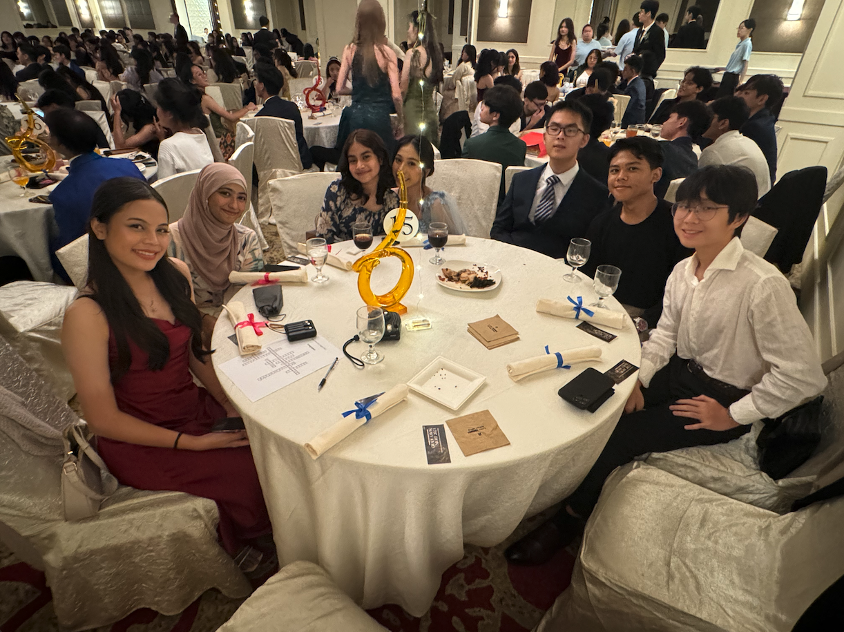Six young people smile at a formal banquet table in a crowded ballroom with white tablecloths and decorations.