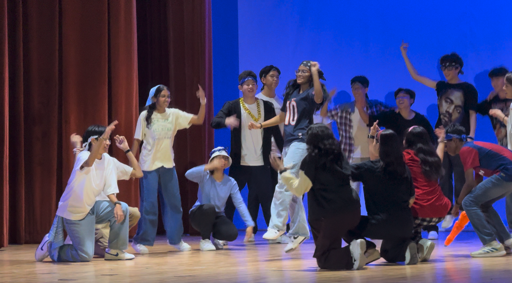 A large group of teens dancing on a stage with red curtains and blue backdrop lighting.
