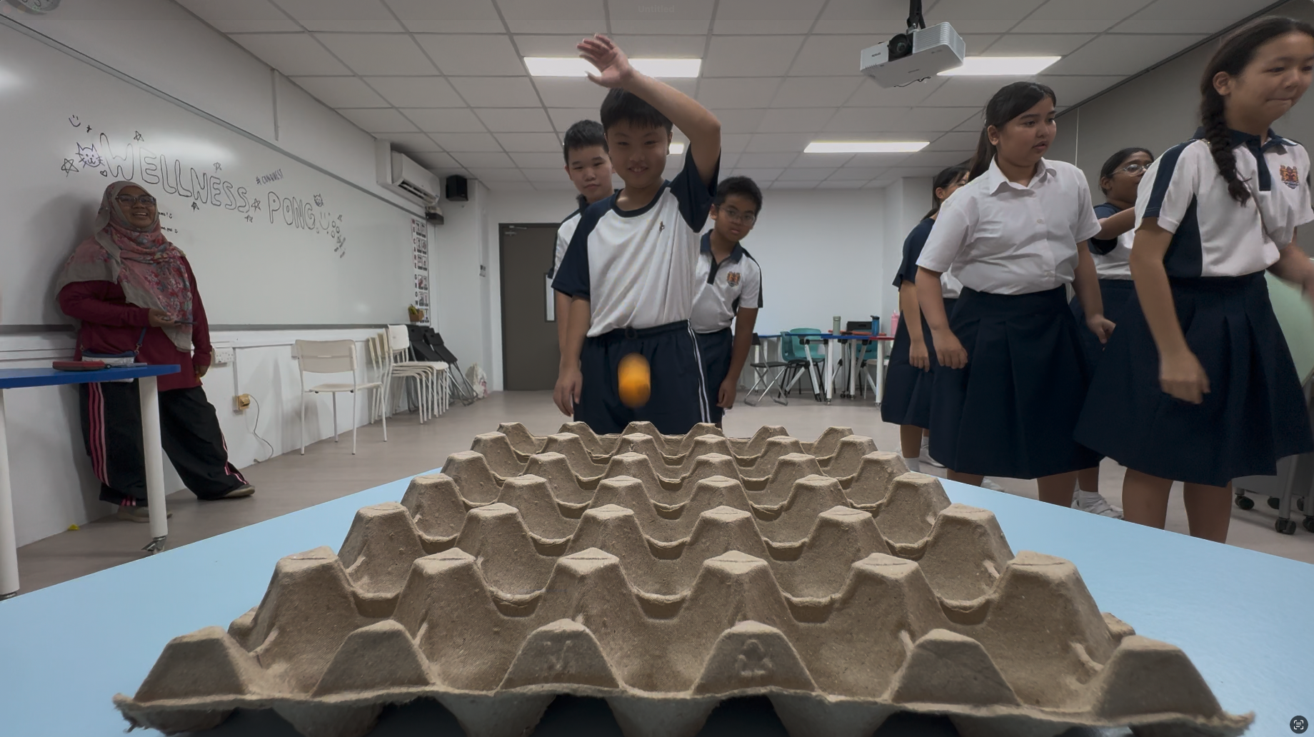 A student tosses an orange ball toward a large egg carton on a blue table during a classroom activity.