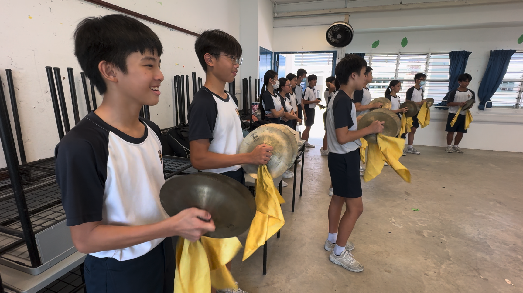 Students in uniforms stand in a row holding metal cymbals with yellow cloths in an indoor hall.