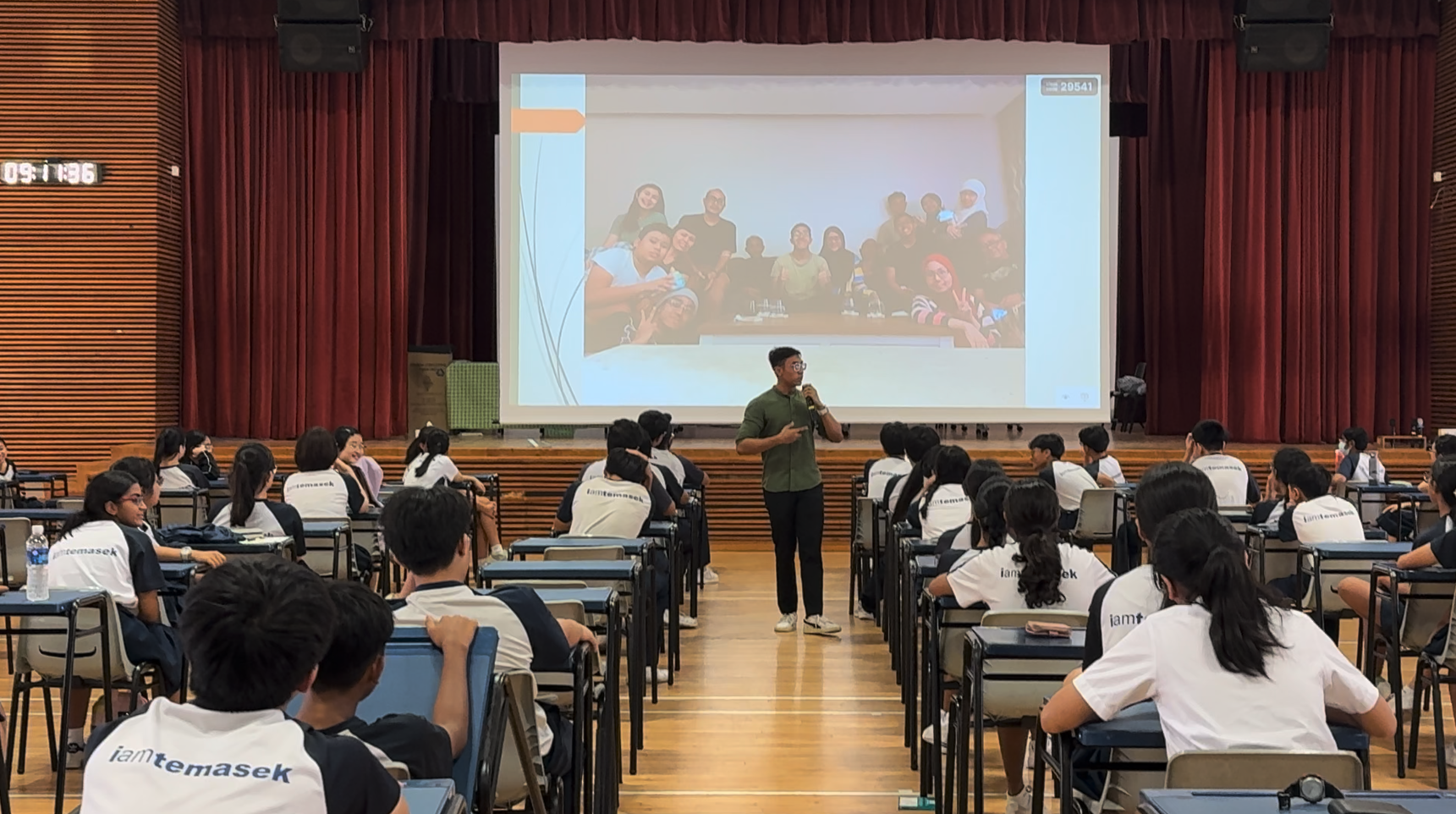A man with a microphone stands before students in white iamtemasek shirts seated in a school hall with a projection screen.