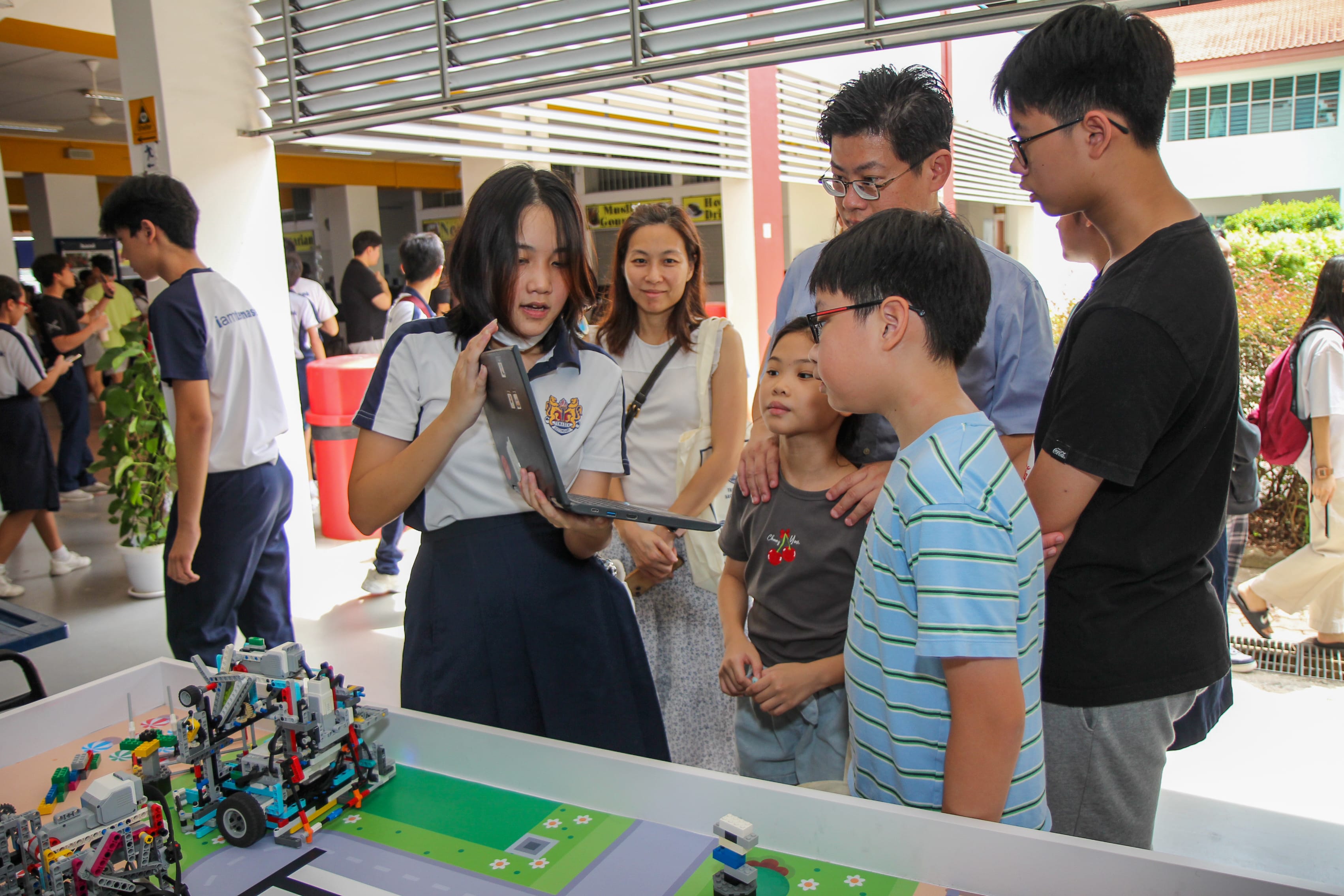 Student in uniform shows a laptop to a small group beside a table with a LEGO robot display.