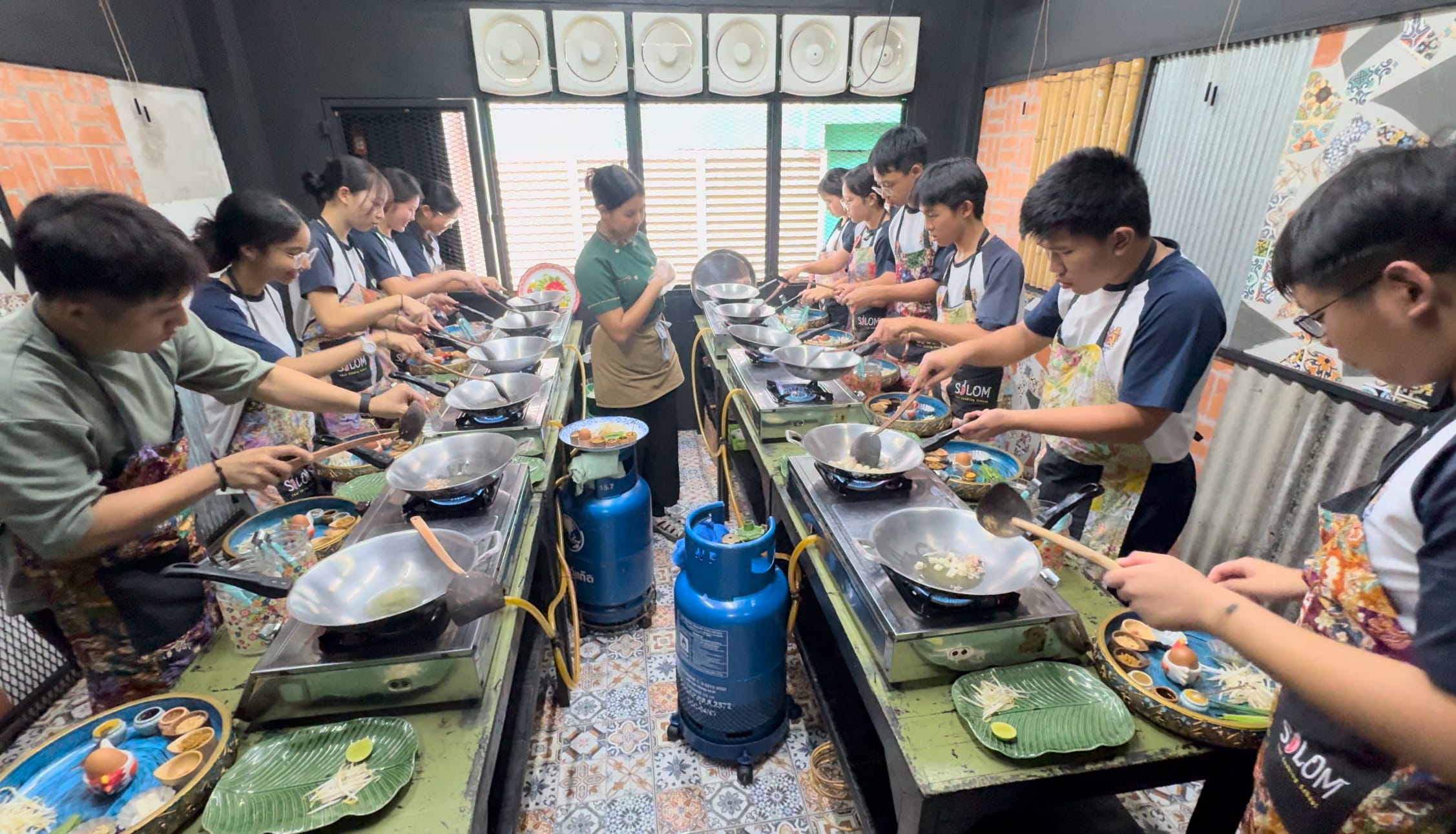 A group of students in aprons cooking at gas stove stations with woks during a Thai cooking class.