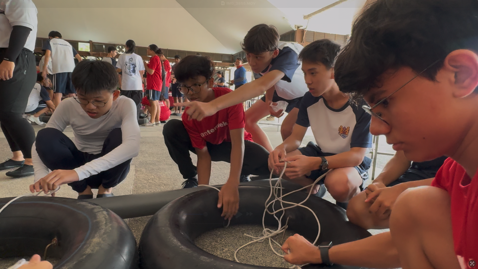 A group of students crouch on the ground working together with rubber tubes and rope during an outdoor team activity.