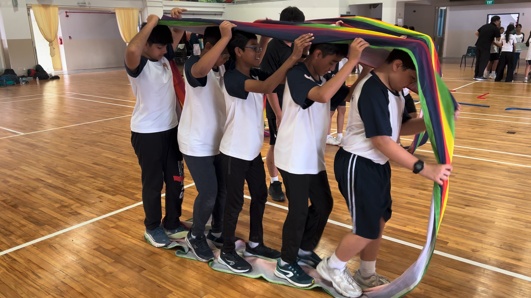 Students in a gymnasium hold a colorful parachute over their heads during a group activity on a wooden floor.