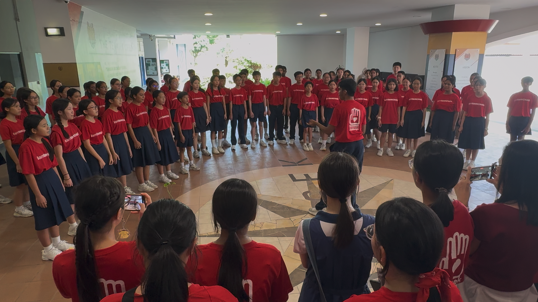 A large student choir in red shirts and navy skirts performs indoors, led by a conductor facing them.