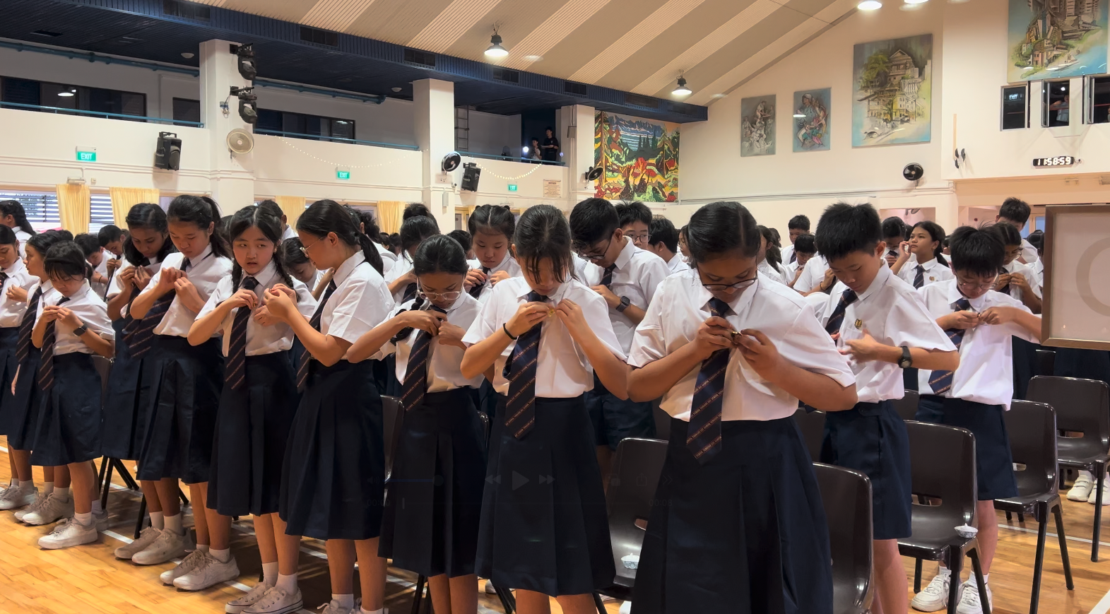 Students in white shirts and navy uniforms pin badges onto their ties in a school hall.