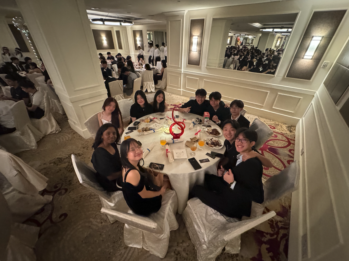 A group of young people in formal black attire seated at a round dining table in an elegant ballroom.