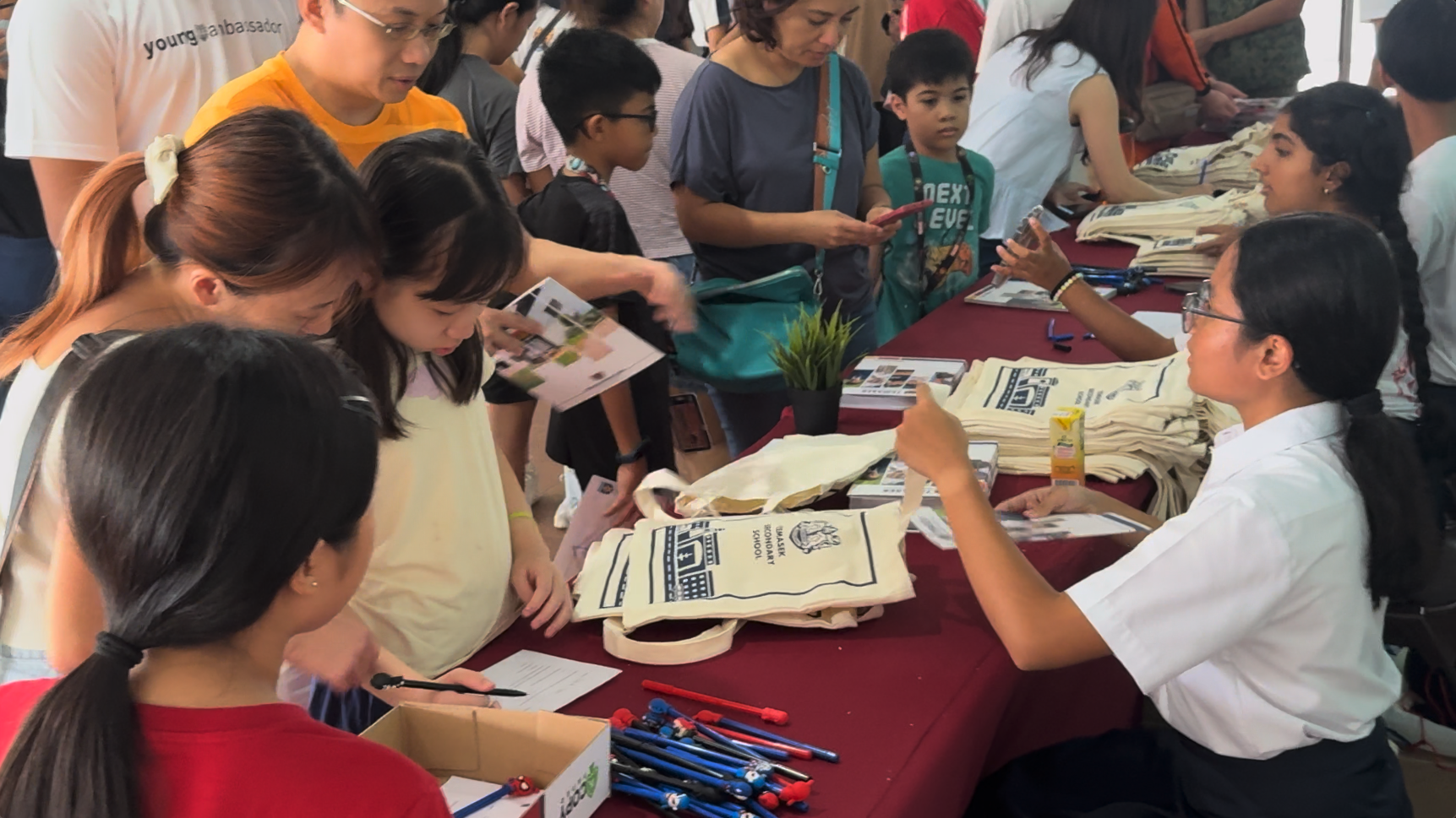 People gathered at a busy event booth with tote bags, brochures, and pens on a red-covered table.