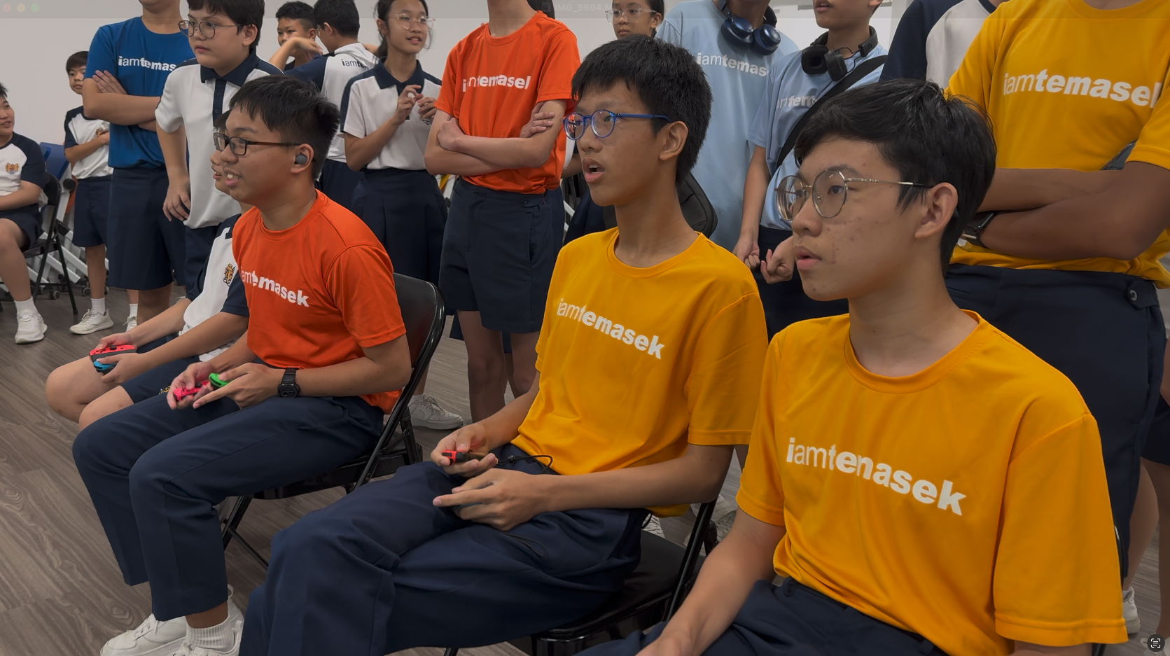 Students in iamtemasek shirts sit holding game controllers while others stand watching in a school room.